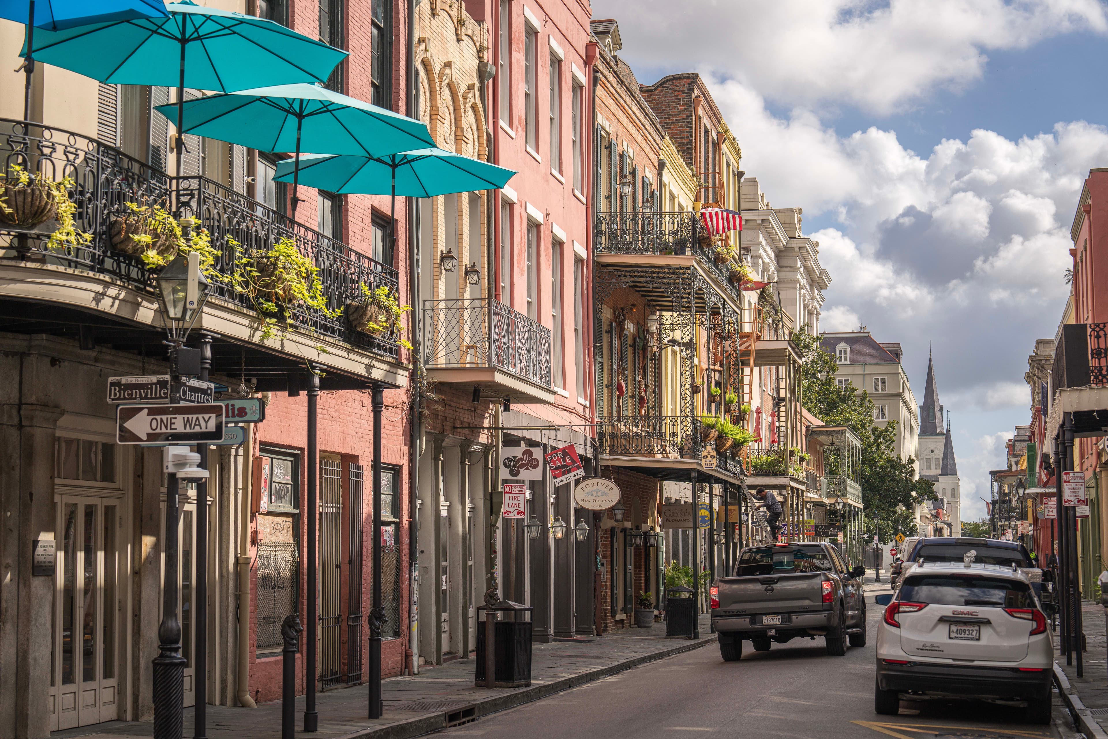 Colorful historic buildings with iron balconies and turquoise umbrellas line a narrow New Orleans street.